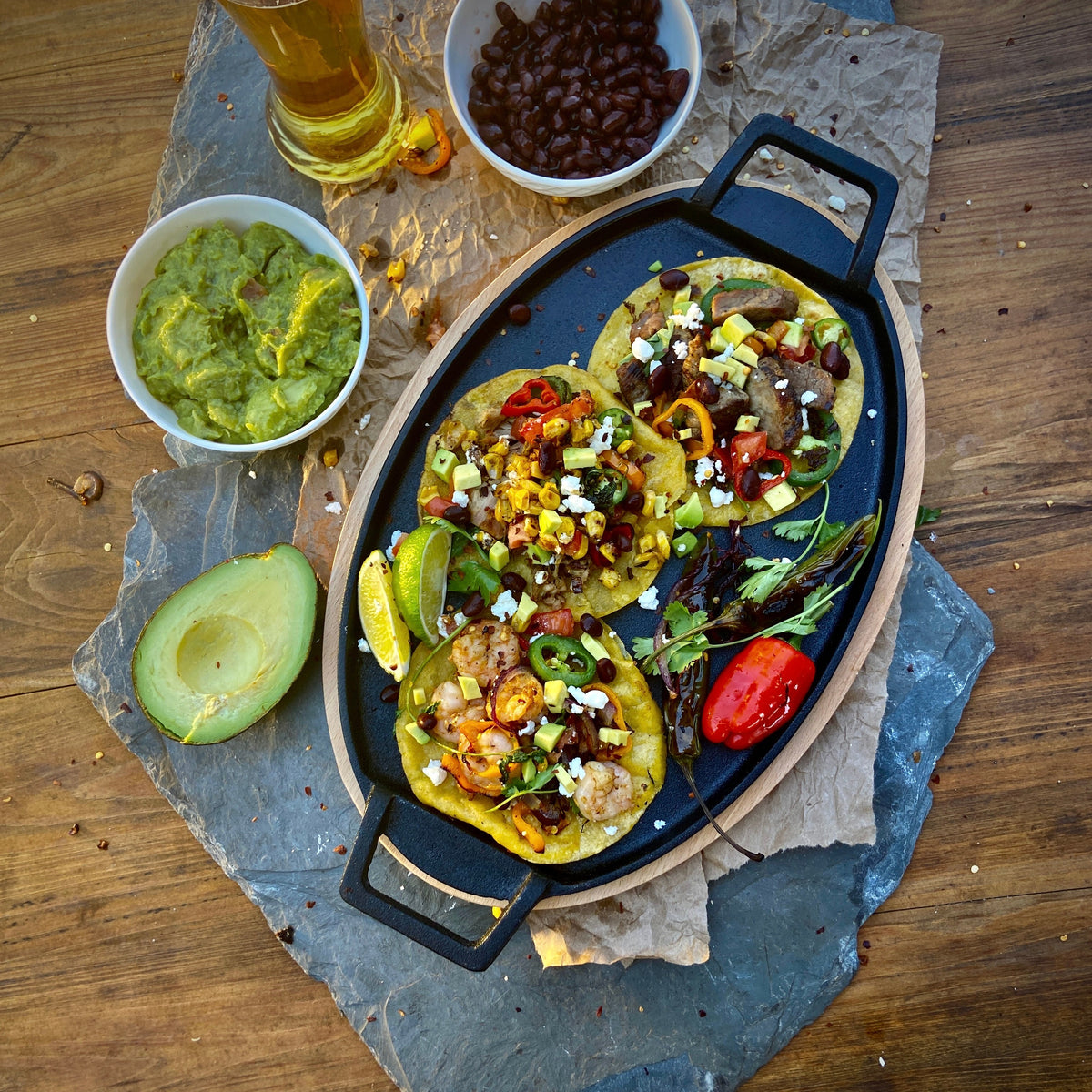 Large cast iron fajita pan on a wooden trivet with three corn street tacos, a bowl of guacamole, and a bowl of black beans.