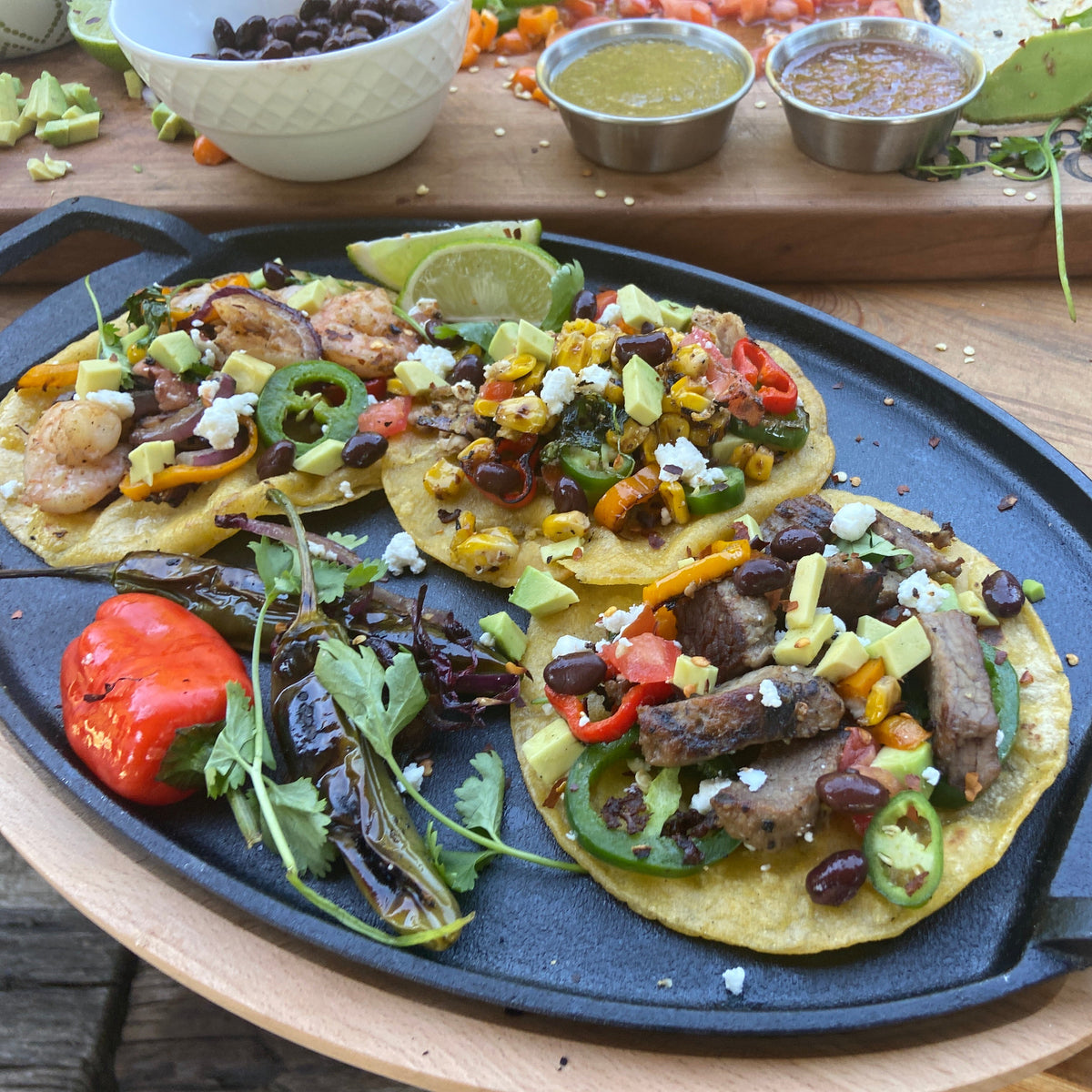 Cast iron fajita pan on a wooden trivet with street tacos on corn tortillas and colorful ingredients.