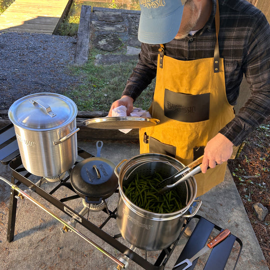 Person outside using triple burner cooker with stainless pot of green beans, an aluminum pot, and a cast iron skillet